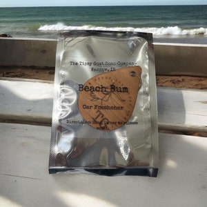 May include: A wooden car freshener shaped like a teardrop with the text "Beach Bum" and "Car Freshener" on it. The freshener is packaged in a silver foil pouch with the text "The Tipsy Goat Soap Company" and "Warsaw, IN".