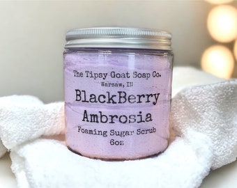 May include: A glass jar with a silver lid filled with a purple sugar scrub. The label reads "The Tipsy Goat Soap Co. Warsaw, Ill Blackberry Ambrosia Foaming Sugar Scrub 6oz"