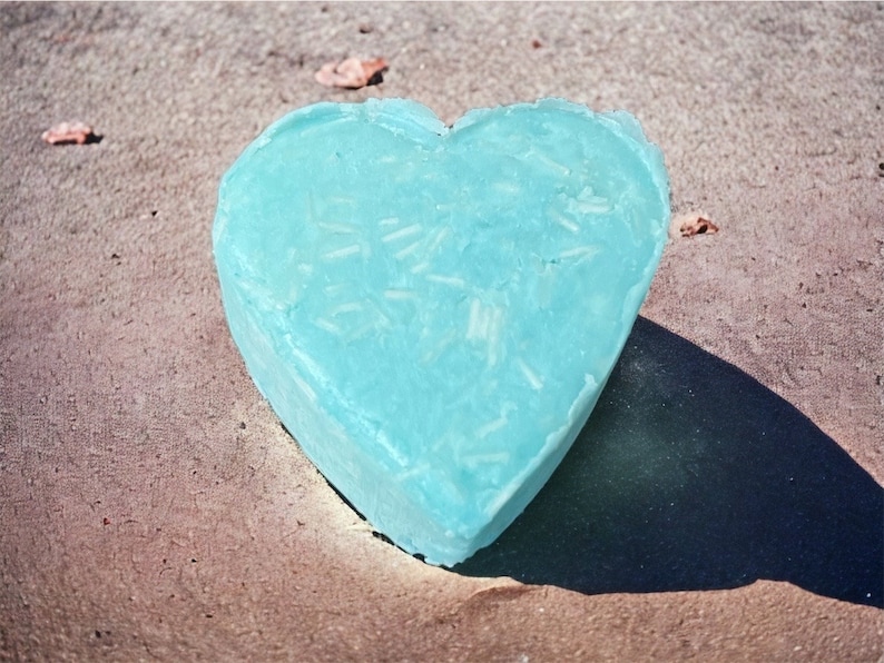 May include: A heart-shaped blue soap bar with white flecks throughout. The soap is sitting on a brown, textured surface.