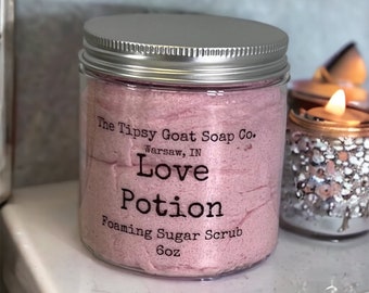 May include: A clear glass jar with a silver lid filled with a pink foaming sugar scrub. The label reads "The Tipsy Goat Soap Co. Warsaw, IN Love Potion Foaming Sugar Scrub 6oz."