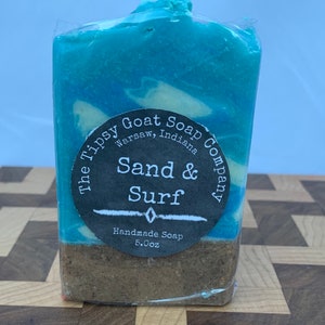 May include: A blue and white swirl handmade soap bar with a black label that reads "The Tipsy Goat Soap Company, Warsaw, Indiana" and "Sand & Surf". The label also states "Handmade Soap 5.0oz".