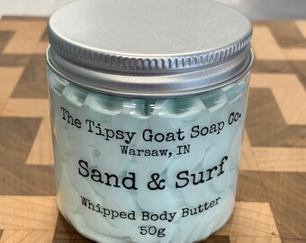 May include: A glass jar with a silver lid filled with a light blue whipped body butter. The jar has a label that reads "The Tipsy Goat Soap Co. Warsaw, IN Sand & Surf Whipped Body Butter 50g".