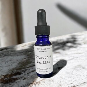 May include: A small blue glass bottle with a black dropper top. The bottle is labeled "Tipsy Goat Soap Co. Warsaw, IN Tobacco & Vanilla Car Freshener Oil 0.5oz (14g)"