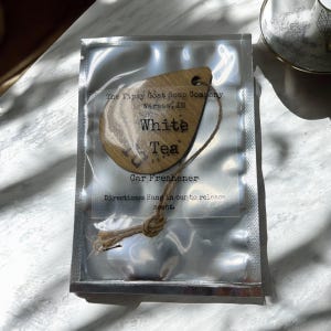May include: A wooden car air freshener with a brown string. The air freshener is shaped like a teardrop and has the text "The Tipsy Goat Soap Company Warsaw, IN White Tea Car Freshener Directions: Hang in car to release scent." printed on it.