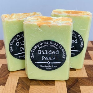 May include: Three bars of green and gold handmade soap with a black label that reads "The Tipsy Goat Soap Company, Warsaw, Indiana, Gilded Pear, Handmade Soap 5.0oz".