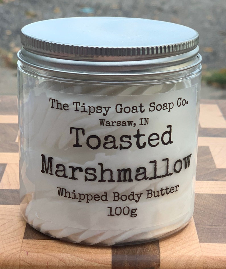 May include: A glass jar with a silver lid filled with white whipped body butter. The label reads "The Tipsy Goat Soap Co. Warsaw, IN Toasted Marshmallow Whipped Body Butter 100g"