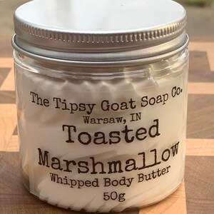 May include: A white jar with a silver lid filled with a white whipped body butter. The label reads "The Tipsy Goat Soap Co. Warsaw, IN Toasted Marshmallow Whipped Body Butter 50g".