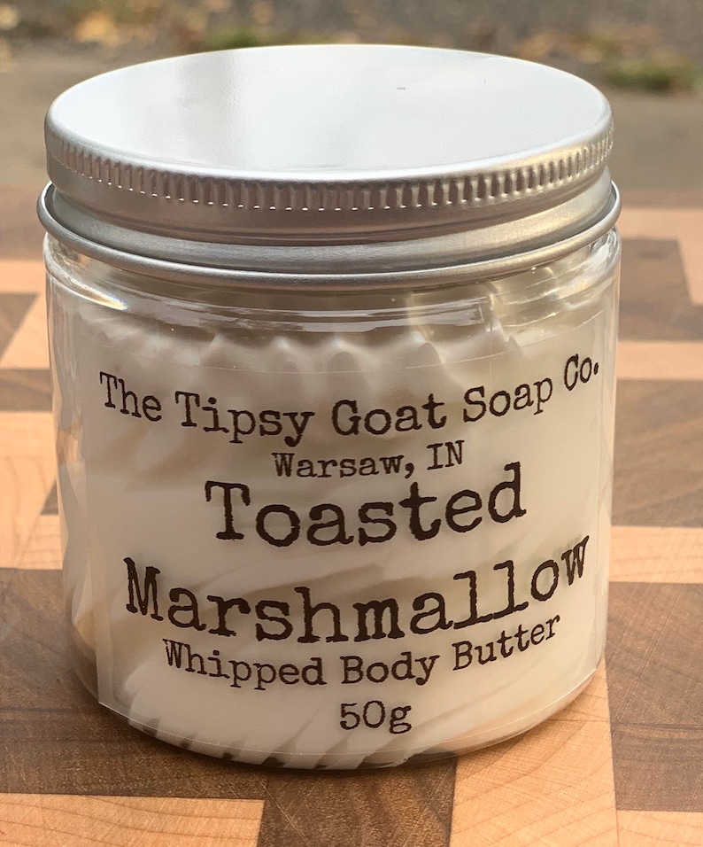 May include: A white jar with a silver lid filled with a white whipped body butter. The label reads "The Tipsy Goat Soap Co. Warsaw, IN Toasted Marshmallow Whipped Body Butter 50g".