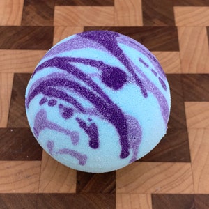 May include: A round, light blue bath bomb with a textured surface. The bath bomb features a swirling pattern of dark purple lines and shapes across its surface. The background is a wooden cutting board.