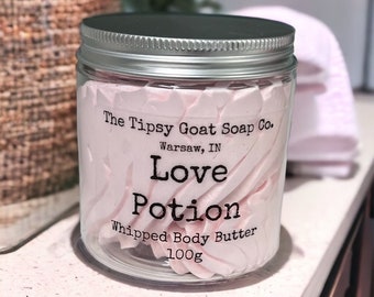 Pode incluir: Um frasco de vidro com tampa prateada cheio de manteiga corporal batida rosa. O rótulo diz "The Tipsy Goat Soap Co. Warsaw, IN Love Potion Whipped Body Butter 100g"