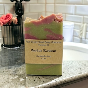 May include: A bar of green and pink handmade soap with a brown label that reads "The Tipsy Goat Soap Company, Warsaw, IN, Cactus Blossom, Handmade Soap, 5.0oz".