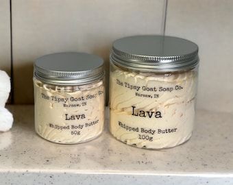Pode incluir: Dois frascos de manteiga corporal batida etiquetados "The Tipsy Goat Soap Co. Warsaw, IN" com o aroma "Lava". O frasco menor é de 50g e o frasco maior é de 100g.