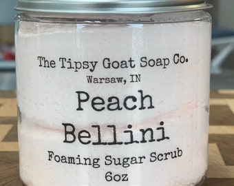 May include: A clear plastic jar with a silver lid filled with a pink foaming sugar scrub. The label reads "The Tipsy Goat Soap Co. Warsaw, IN Peach Bellini Foaming Sugar Scrub 6oz."
