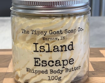 May include: A clear jar with a silver lid filled with a white whipped body butter. The label reads "The Tipsy Goat Soap Co. Warsaw, IN Island Escape Whipped Body Butter 100g".