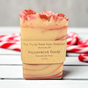 May include: A rectangular bar of handmade soap with a Peppermint Candy scent. The soap is beige with red swirls and topped with red chunks. A brown paper band wraps around the soap, displaying the company name, location, and product details. The soap weighs 5.0 oz.