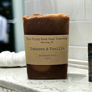 May include: A rectangular bar of handmade soap with a brown and tan color gradient. The label reads "The Tipsy Goat Soap Company, Tobacco & Vanilla, Handmade Soap, 5.0oz."