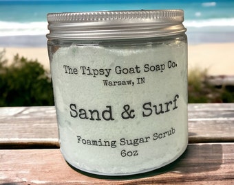 May include: A glass jar with a silver lid filled with a light blue foaming sugar scrub. The jar has a label that reads "The Tipsy Goat Soap Co. Warsaw, IN Sand & Surf Foaming Sugar Scrub 6oz".