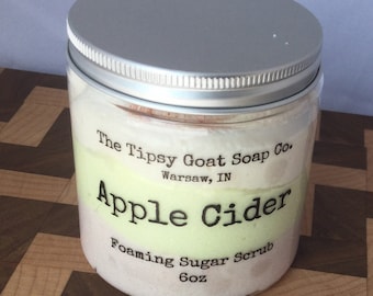 Op de afbeelding: Een glazen pot met een zilveren deksel gevuld met een witte en groene schuimende suiker scrub. Het etiket vermeldt "The Tipsy Goat Soap Co. Warsaw, IN Apple Cider Foaming Sugar Scrub 6oz"