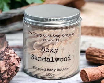 Pode incluir: Um frasco de vidro com tampa prateada cheio de manteiga corporal batida de cor marrom claro. O frasco tem um rótulo que diz "The Tipsy Goat Soap Company, Warsaw, IN, Sexy Sandalwood, Whipped Body Butter, 100g".