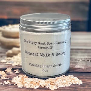 May include: A glass jar with a silver lid containing a white foaming sugar scrub. The label reads "The Tipsy Goat Soap Company, Warsaw, IN, Oatmeal Milk & Honey, Foaming Sugar Scrub, 6oz."