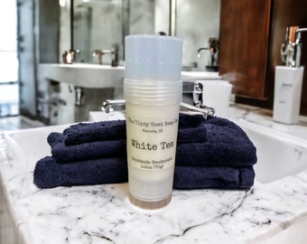 May include: A white plastic deodorant stick with a label that reads "The Tipsy Goat Soap Co. Warsaw, IN White Tea Handmade Deodorant 2.5oz (70g)". The stick is sitting on a stack of dark blue towels.