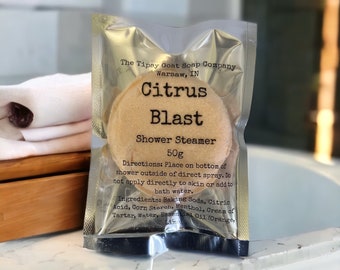May include: A white pouch with a round, beige shower steamer inside. The pouch has black text that reads "The Tipsy Goat Soap Company, Warsaw, IN, Citrus Blast, Shower Steamer, 50g". The text also lists the ingredients: Baking Soda, Citric Acid, Corn Starch, Menthol, Cream of Tartar, Water, Essential Oil (Orange).