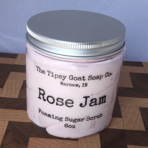 May include: A glass jar with a silver lid filled with a pink foaming sugar scrub. The label reads "The Tipsy Goat Soap Co. Warsaw, IN Rose Jam Foaming Sugar Scrub 6oz"