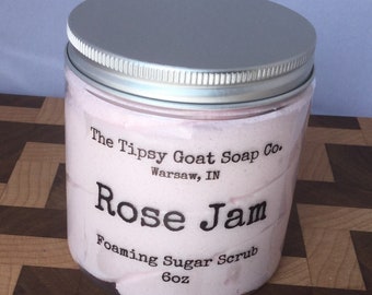 Op de afbeelding: Een glazen pot met een zilveren deksel gevuld met een roze schuimende suiker scrub. Het etiket luidt "The Tipsy Goat Soap Co. Warsaw, IN Rose Jam Foaming Sugar Scrub 6oz"