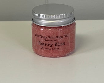 Op de afbeelding: Een klein roze potje met een zilveren deksel. Het potje bevat een lippen scrub en heeft een label met de tekst "The Tipsy Goat Soap Co. Warsaw, IN Cherry Kiss Lip Scrub 1.25oz."