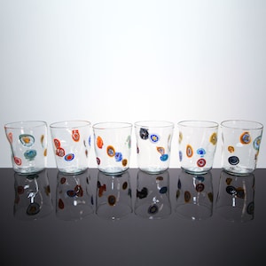 Könnte beinhalten: Sechs klare Trinkgläser mit farbenfrohen, abstrakten Designs. Jedes Glas zeigt eine einzigartige Anordnung von roten, blauen, gelben und orangenen kreisförmigen Mustern. Die Gläser sind in einer Reihe angeordnet und spiegeln sich auf einer dunklen Oberfläche.
