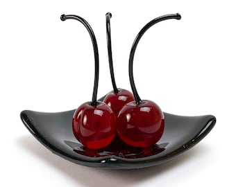 May include: Three red glass cherries with black stems on a black square plate.