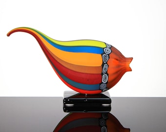 May include: A colorful glass sculpture with a curved, elongated shape. The sculpture features bands of red, orange, yellow, blue, and brown. The design includes black and white circular patterns. It rests on a black rectangular base.
