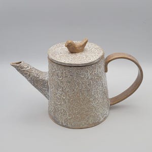May include: A ceramic teapot with a lid and handle. The teapot is beige with a floral pattern and a bird-shaped knob on the lid. The handle and spout are a darker beige color.
