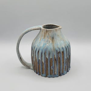 May include: A handmade ceramic pitcher with a large handle. The pitcher has a textured, ribbed design on the lower portion and is glazed in shades of blue and brown, with a dripped effect. The opening is wide and the handle is curved.