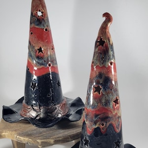 May include: Two ceramic witch hats with pointed, curved tops and wide, wavy brims. The hats feature a glossy glaze in black, red, and blue, with star-shaped cutouts. One hat has a spider detail on the band, while the other features a skull.