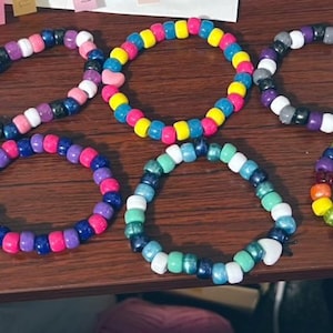Könnte beinhalten: Eine Sammlung von zehn bunten Freundschaftsarmbändern aus kleinen Plastikperlen. Die Armbänder sind in verschiedenen Farben, darunter rosa, blau, gelb, grün, lila, rot, orange und schwarz. Einige der Armbänder haben einen Herz-Anhänger.