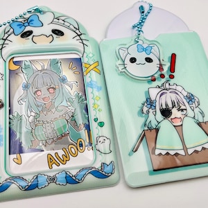 May include: Two light green and blue anime-style card holders with matching keychains. One features a cartoon character with the word "Awoo!" and the other has a character peeking out of a box. Both have a cat-shaped design at the top.