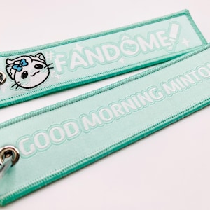 Pode incluir: Dois chaveiros de tecido verde-claro com texto branco e um rosto de gato de desenho animado. Um chaveiro diz "#FANDOME!" e o outro diz "GOOD MORNING MINTO!". Ambos têm uma borda verde e argolas prateadas.