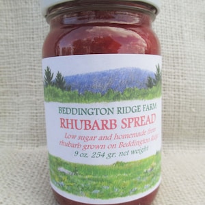 May include: A jar of rhubarb spread with a white lid and a label featuring a landscape illustration. The label reads "Beddington Ridge Farm Rhubarb Spread" and includes the text "9 oz. net weight". The jar is filled with a reddish-pink preserve.
