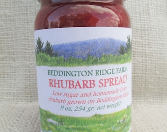 May include: A jar of rhubarb spread with a white lid and a label featuring a landscape illustration. The label reads "Beddington Ridge Farm Rhubarb Spread" and includes the text "9 oz. net weight". The jar is filled with a reddish-pink preserve.