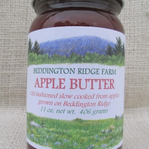 May include: A jar of apple butter with a white lid. The label reads "Beddington Ridge Farm Apple Butter" with a landscape illustration. The jar contains 11 oz. net wt. or 406 grams of apple butter.