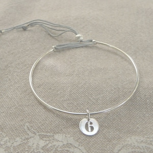 May include: A silver bangle bracelet with a silver charm that has the number 6 on it. The bracelet is tied with a gray cord.