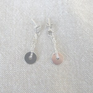 May include: A pair of silver drop earrings. Each earring features a small, round disc suspended from a delicate chain. The chain connects to a post-style earring back. The earrings are displayed against a neutral, textured background.