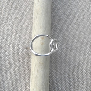 Peut inclure: Une bague en argent avec un design circulaire. La bague présente un petit cercle connecté d'un côté du cercle plus grand. La bague est présentée sur une cheville en bois sur un fond neutre.