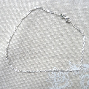 Puede incluir: Una delicada pulsera de cadena plateada con un diseño de cuerda trenzada. La pulsera está colocada sobre una tela beige claro. El cierre es visible en un extremo, y la cadena parece estar hecha de pequeños eslabones interconectados.