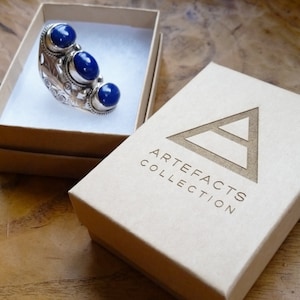 Peut inclure: Une bague en argent avec trois pierres pr&eacute;cieuses bleues serties dans un motif floral. La bague est dans une petite bo&icirc;te blanche avec les mots "Artefacts Collection" imprim&eacute;s sur le c&ocirc;t&eacute;.