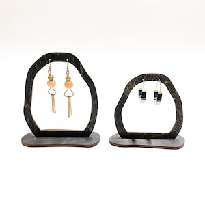 May include: Two wooden earring stands with a dark wood finish. One stand has a pair of gold earrings with peach-colored beads and gold tassels. The other stand has a pair of blue and gold hoop earrings.