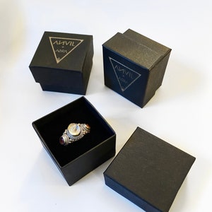 May include: Three black boxes with the text "ANVIL + AURA" on the top. One box contains a silver ring with three stones.