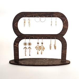 May include: A dark brown wooden earring stand with two tiers and multiple hooks for displaying earrings. The stand is shaped like a figure eight and has a smooth, polished finish.