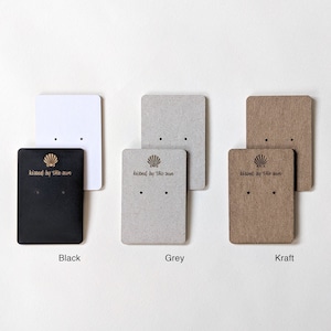 May include: Three earring display cards with a seashell and the text "kissed by the sun". The cards are black, grey, and kraft paper.